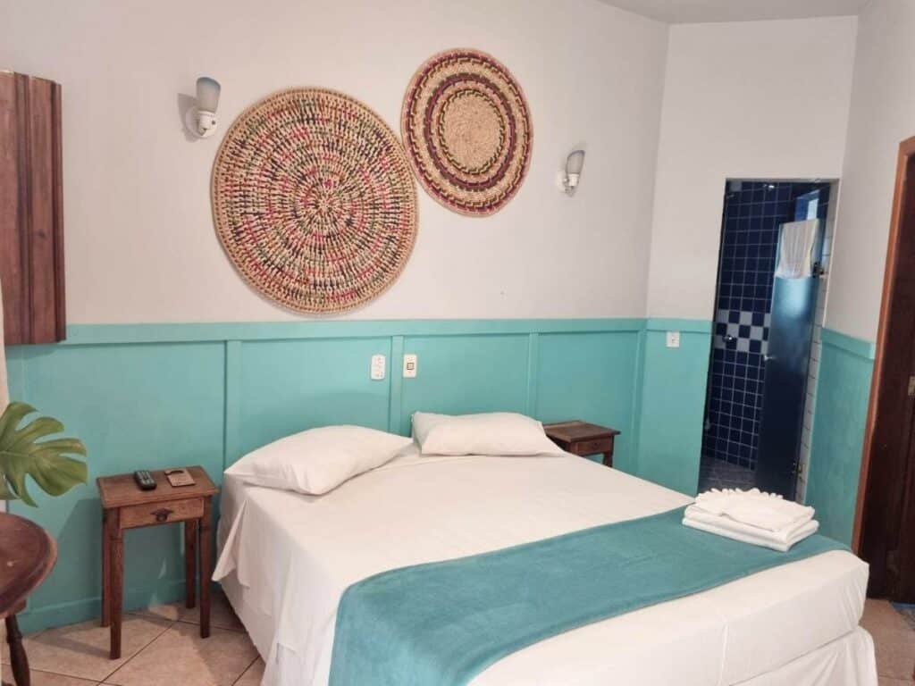 Quarto no VELINN Hotel Santa Tereza com uma cama de casal, duas mesinhas de madeira, tudo decorado em azul e madeira, para representar resorts em Ilhabela