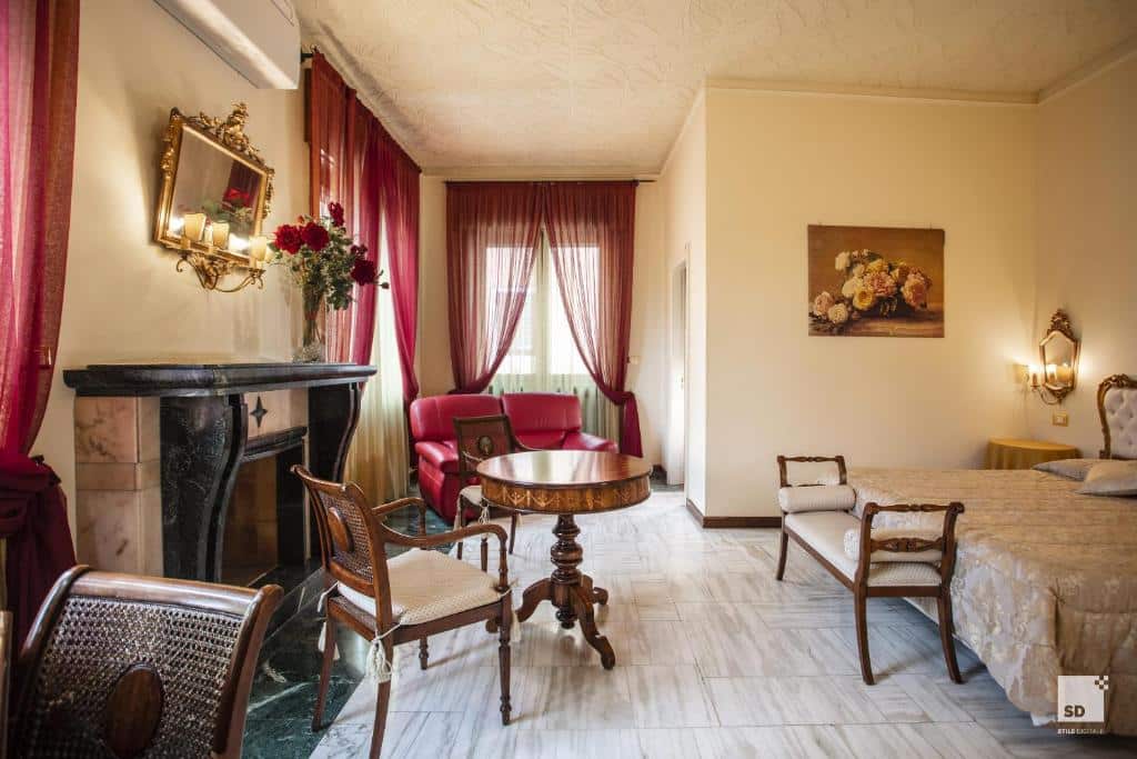 quarto do Hotel Alessandro Della Spina, um dos hotéis em Pisa, com cama de casal grande, mesa com cadeiras e poltronas em frente a uma lareira e ainda um sofá do outro lado com janelas e cortinas vermelhas