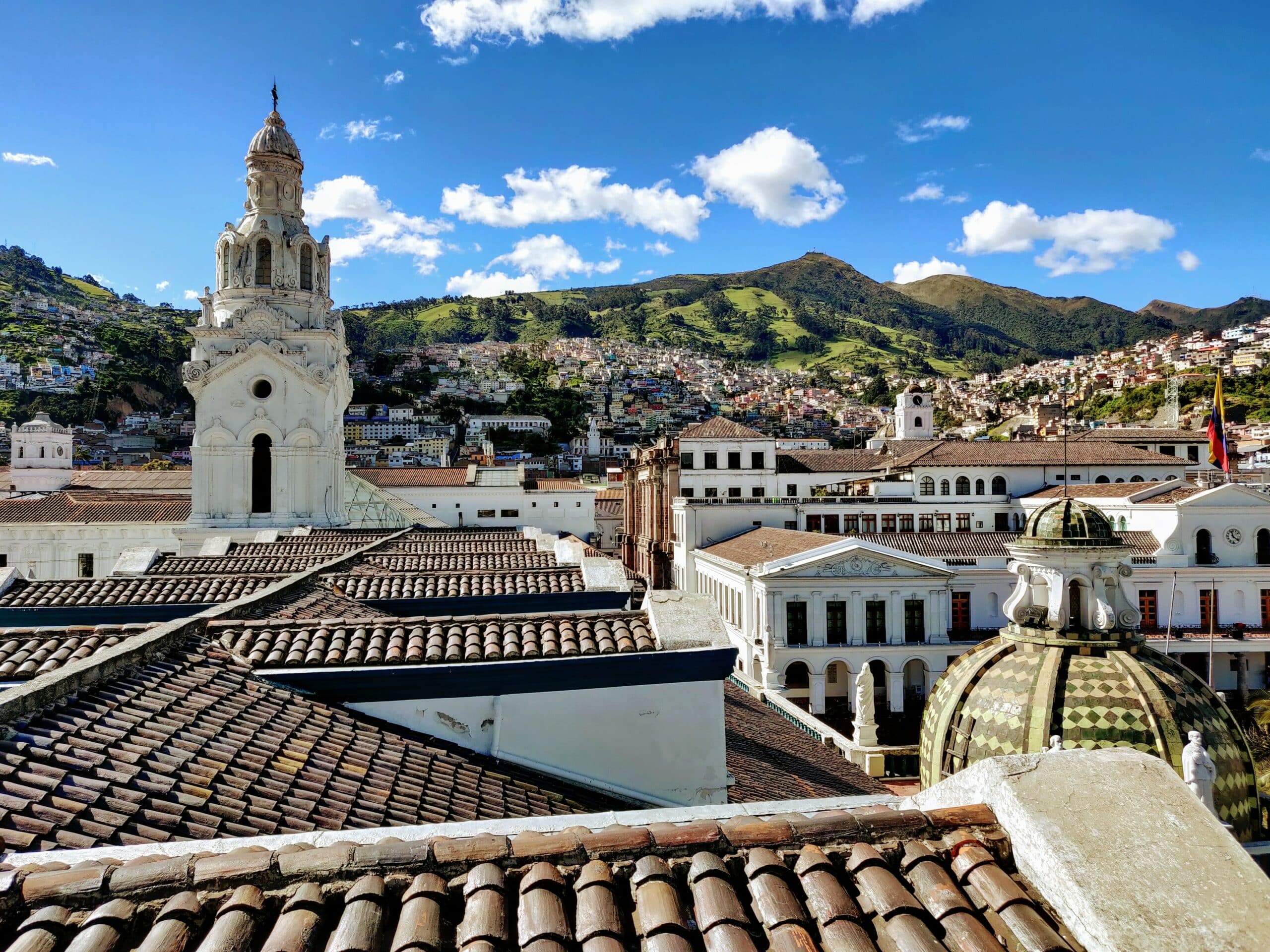Vista da cidade de Quito, Equador com várias casas, igreja, ao fundo montanhas verdes com céu claro.
