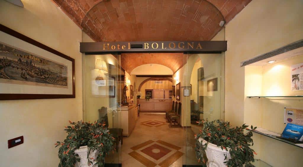 recepção do Hotel Bologna, um dos hotéis em Pisa, com quadros dos lados, entrada de vidro e o balcão de um lado e um corredor do outro