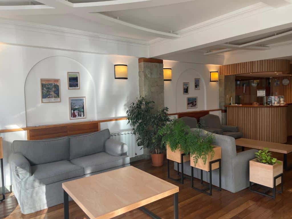 Recepção do Hotel Ayres Del Nahuel com sofá de dois lugares cinza, mesa de madeira a frente, do lado esquerdo da imagem tem dois vasos de plantas, um sofá cinza e duas poltronas com mesa de centro. Representa hotéis em Bariloche.