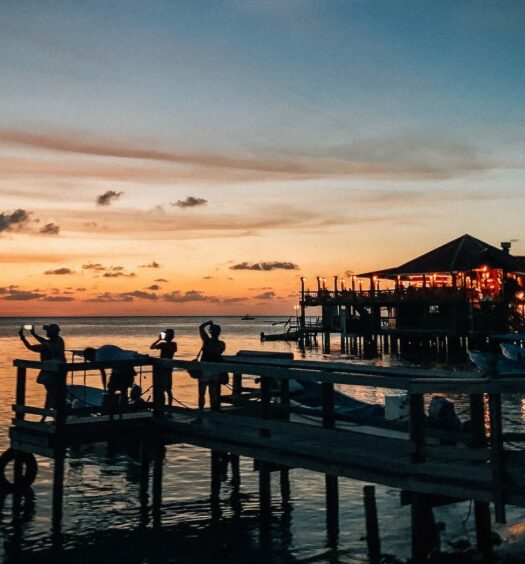 vista do pôr do sol da praia de Roatán, com pessoas tirando foto a partir de um cais de madeira que se estende até a água com um bangalô ao fundo mais afastado, para ilustrar o post de chip celular Honduras