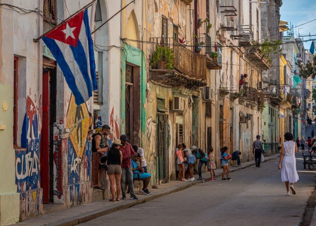 Viela de Havana com algumas pessoas andando e conversando, além de ter casas antigas, sendo uma com a bandeira de Cuba pendurada, e uma calçada curta com alguns moradores sentados