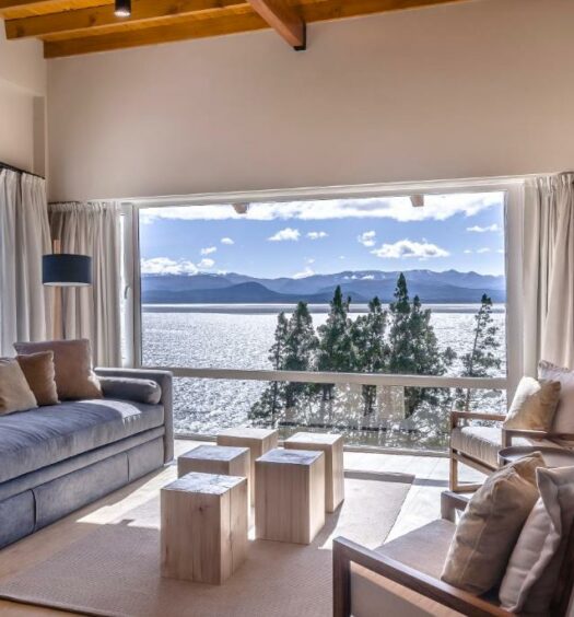 Sala de estar do Aguila Mora Suites & Spa com sofá cinza, bancos de madeira com mesas de centro, duas poltronas em frente ao sofá e ao fundo há uma janela panorâmica com vista para a praia. Para ilustrar os hotéis em Bariloche