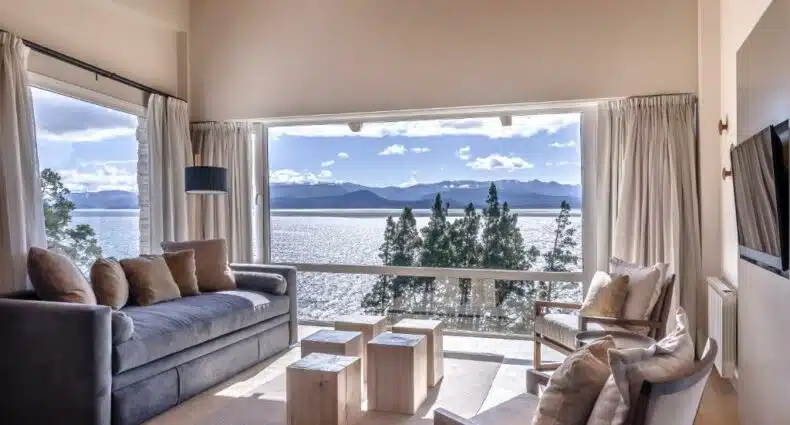 Sala de estar do Aguila Mora Suites & Spa com sofá cinza, bancos de madeira com mesas de centro, duas poltronas em frente ao sofá e ao fundo há uma janela panorâmica com vista para a praia. Para ilustrar os hotéis em Bariloche