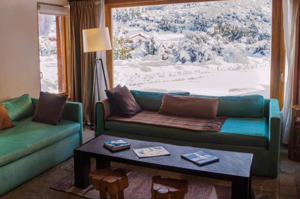 Sala do Galileo Boutique Hotel com dois sofás verdes com almofadas marrom em cima, mesa de centro de madeira com dois bancos rústicos e ao fundo janela ampla com vista para fora com a neve cobrindo tudo. Representa hotéis em Bariloche