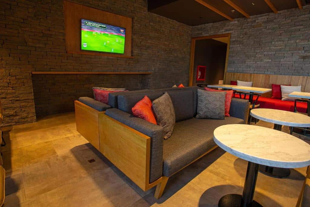 Sala do Hampton By Hilton Bariloche com sofá preto, mesas de mármores e ao fundo  uma TV na parede de tijolos. Representa hotéis em Bariloche.