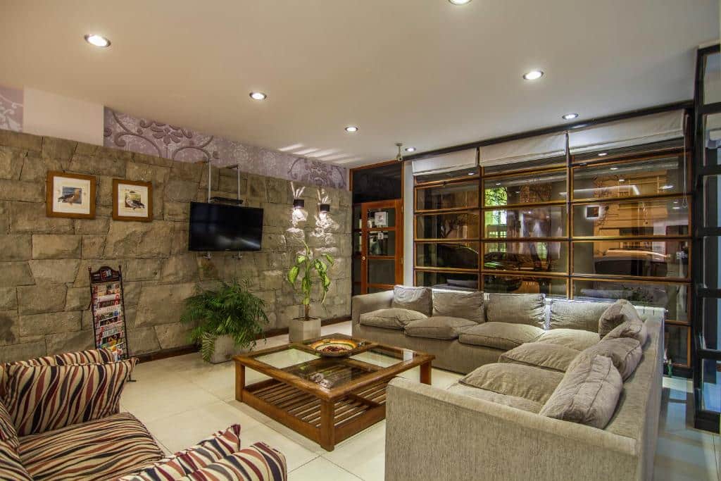 Sala de estar do Hotel Milan com sofá de cor cinza, mesa de centro de madeira, sofá listrado de azul e vermelho do lado direito em frente a mesa, TV na parede de pedra com dois vasos de plantas a frente. Representa hotéis em Bariloche.