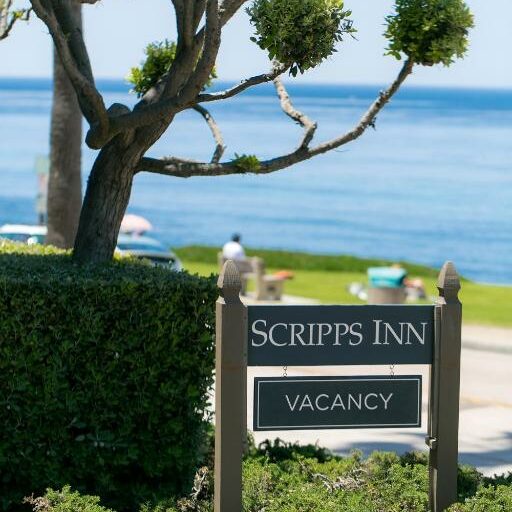Placa escrito "Scripps Inn Vacancy", com uma árvore ao lado, e vista para a praia em um dia ensolarado ao fundo