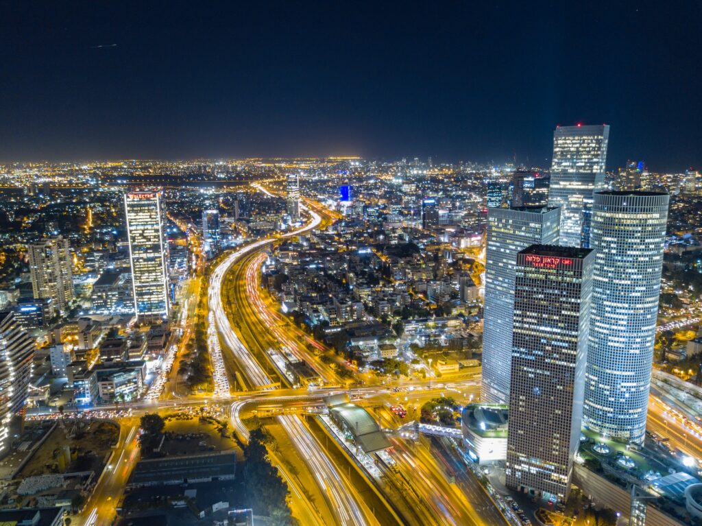 Foto aérea da cidade de Tel Aviv com prédios e arranha-céus modernos e iluminados, via com carros circulando em uma noite na cidade para representar o seguro viagem Tel Aviv.