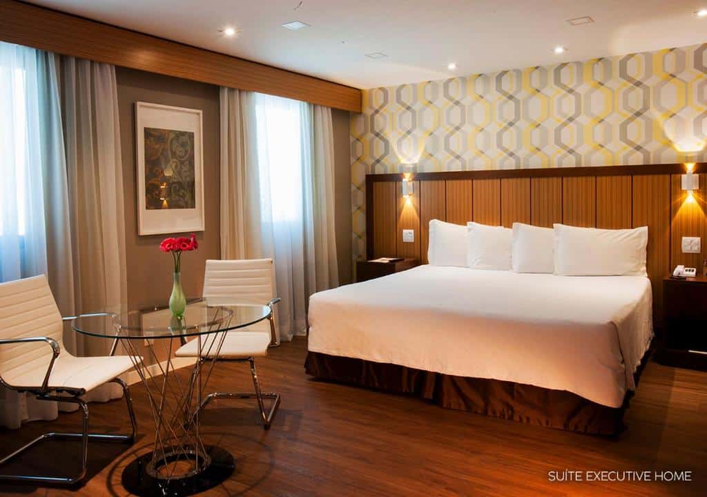 Quarto no Bourbon São Paulo Ibirapuera Convention Hotel com uma cama de casal com muitos travesseiros, chão de madeira, uma pequena mesinha redonda de vidro com duas cadeiras, duas janelas com cortinas, e uma cabeceira com iluminação indireta