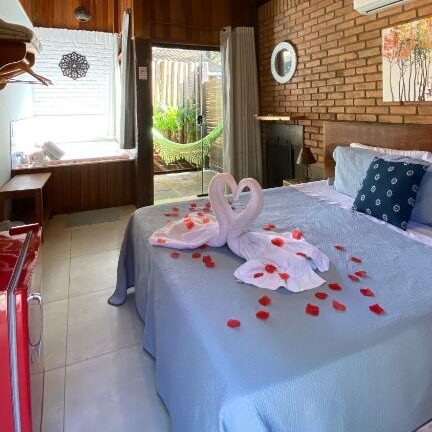 Suíte deluxe, de 27 m², de uma das pousadas na Serra da Mantiqueira (Xai Mauá), com uma cama de casal com folhas vermelhas e uma escultura feita de toalhas brancas em cima, além de uma banheira de hidromassagem iluminada e uma varanda pequena com uma rede  verde limão