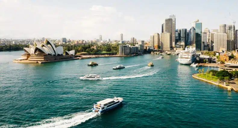 Vista aérea do Porto de Sydney com a Ópera, barcos na água e arranha-céus da cidade em um dia ensolarado. A imagem ilustra o post sobre intercâmbio na Austrália.
