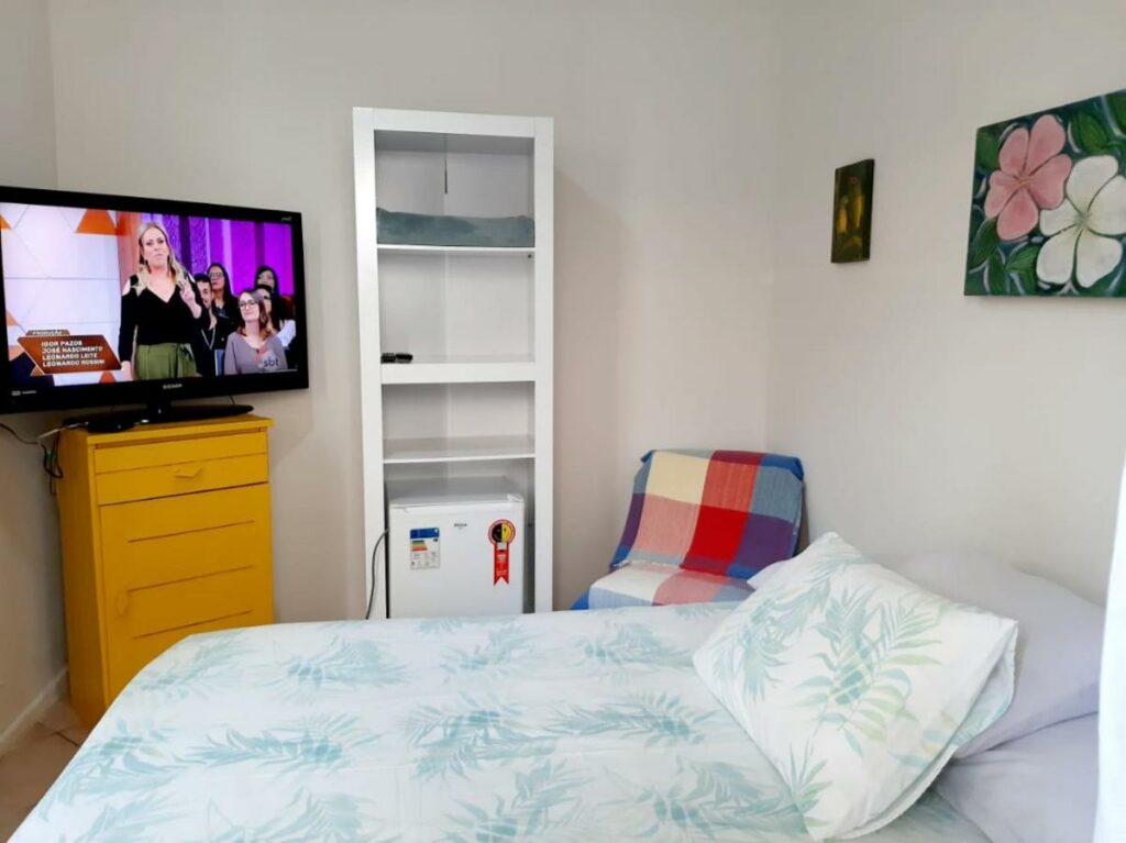 Suíte do Tartaruga Marinha Guest House com uma pequena estante, uma cômoda com uma televisão, um frigobar, uma cama de casal e uma cadeira
