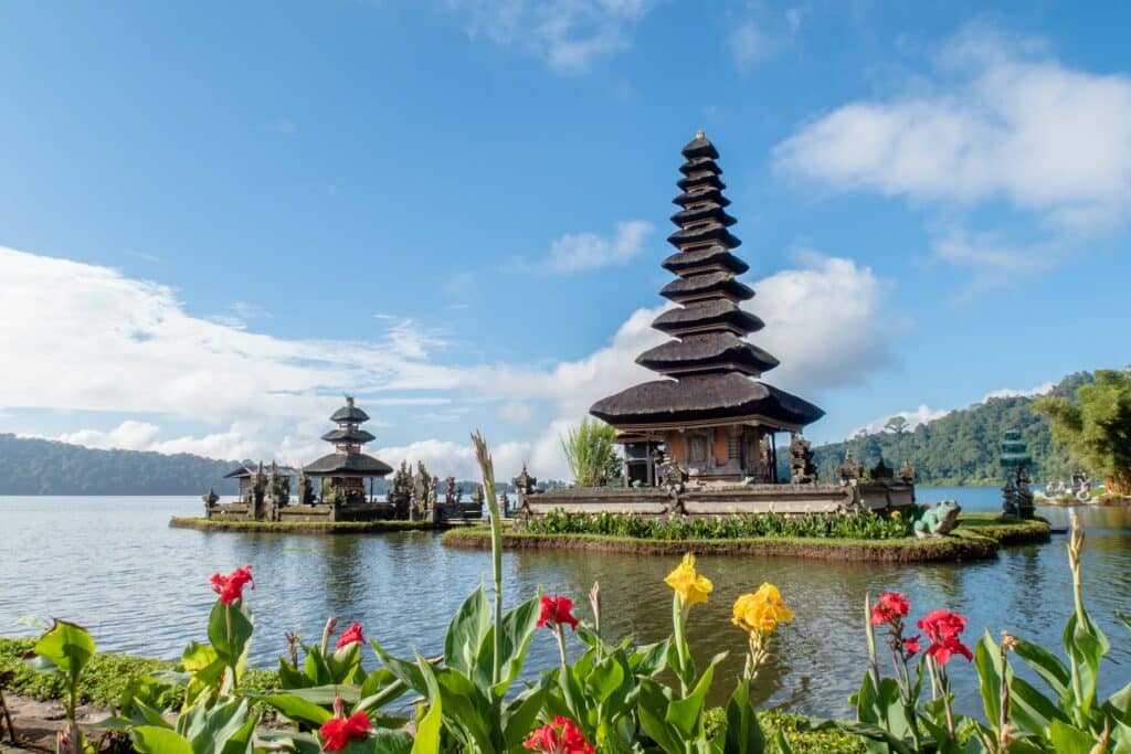 vista do Ulun Danu Beratan, que é uma estrutura bem alta em forma de cone, a base do telhado é mais larga e a ponta, mais compacta, há um lado nos arredores e flores coloridas amarelas, rosas e laranjas emoldurando o foto com um céu azul com nuvens, para ilustrar o post de chip celular Bali