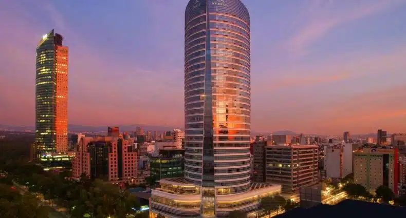 Prédio alto e espelhado do The St Regis Mexico City, um dos hotéis na Cidade do México. no início da noite com o pôr do sol amarelado refletindo no prédio, além da vista da rua cercada por árvores onde fica o local e vários outros prédios ao redor