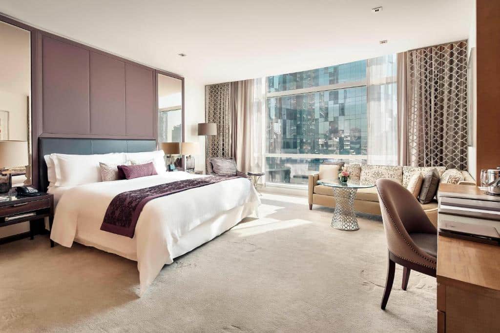 Quarto espaçoso do hotel The St Regis, com cama de casal, sofá, mesa com poltrona e uma parede de vidro ampla com vista de um prédio espelhado
