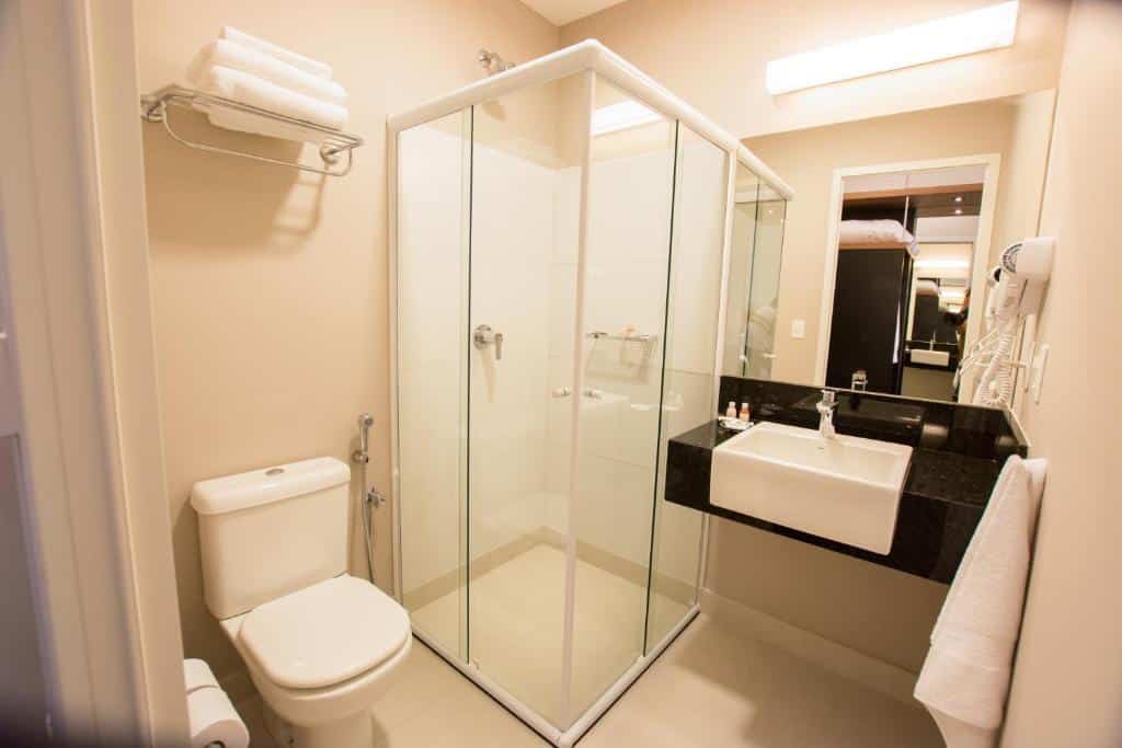 Banheiro do Travel Inn Wise Indaiatuba com box de vidro, vaso sanitário e uma pia com espelho, para representar os hotéis perto do Aeroporto de Viracopos em Campinas