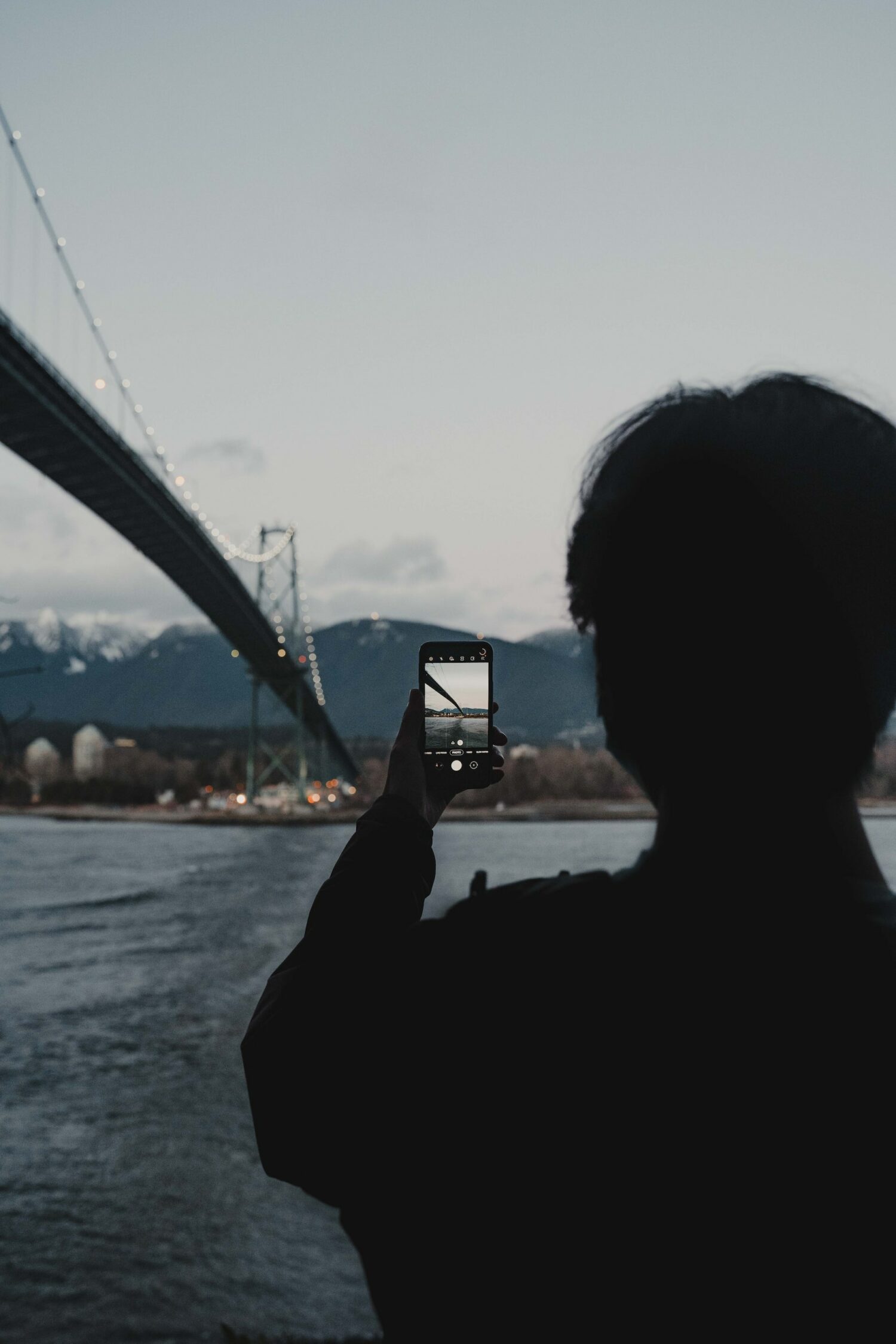 Silhueta de uma pessoa fotografando a ponte de Vancouver utilizando um celular em um dia nublado com algumas nuvens.