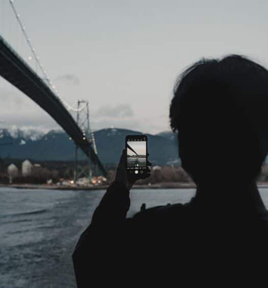 Silhueta de uma pessoa fotografando a ponte de Vancouver utilizando um celular em um dia nublado com algumas nuvens.