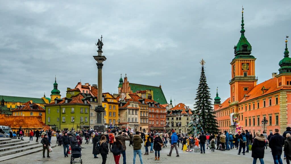 Várias pessoas andando em uma praça da Varsóvia, capital da Polônia, durante um dia nublado, com várias construções históricas em volta e uma árvore de natal grande e alta no meio