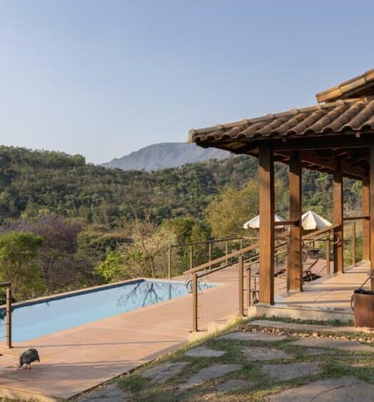 vista da serra da Villa Severina, uma das pousadas em Brumadinho, com a entrada de um lado com detalhes em madeira e do outro, uma piscina retangular comprida que dá vista para a vegetação e a serra