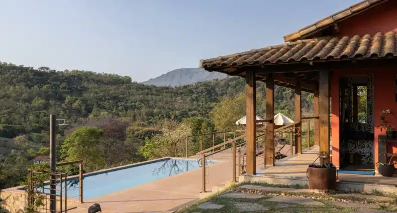 vista da serra da Villa Severina, uma das pousadas em Brumadinho, com a entrada de um lado com detalhes em madeira e do outro, uma piscina retangular comprida que dá vista para a vegetação e a serra