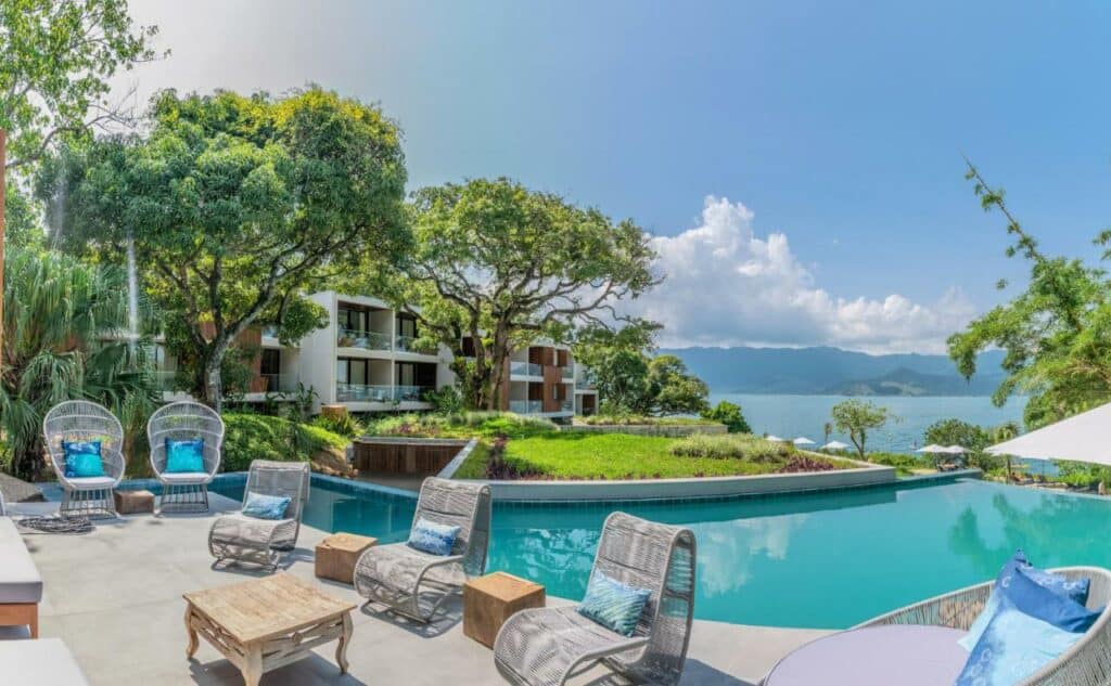 Uma piscina no Wyndham Ilhabela Casa Di Sirena com borda infinita em direção do mar e algumas poltronas pra se sentar no deck.