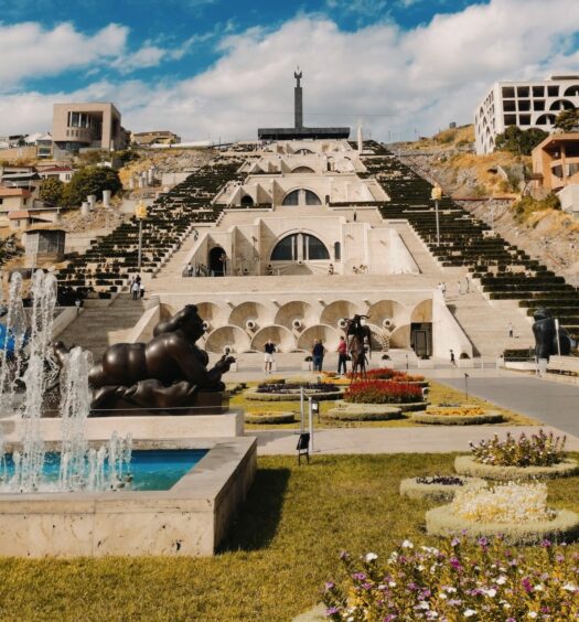 praça com uma longa escadaria que forma quase uma pirâmide, em Yerevan, capital da Armênia, que ilustra o post de chip celular Leste Europeu, com uma fonte e escultura de pessoa toda em preto, pessoas passeiam pela praça e um homem tira uma foto com o celular da cena