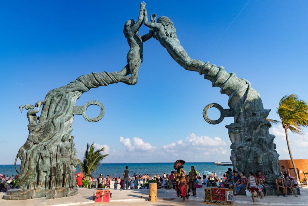 Duas estátuas grandes de mulheres com uma espécie de calda de sereia bem en frente a entrada de uma praia cheia de pessoas passando perto