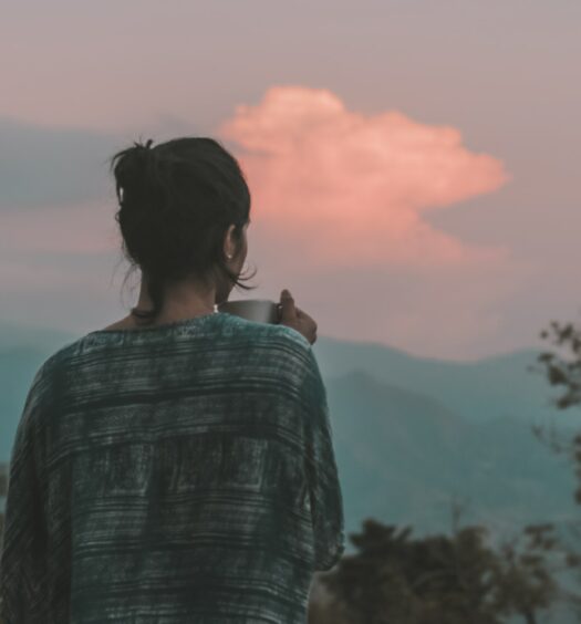 mulher de costas com uma caneca na mão observando um vale com árvores e montanhas ao nascer do sol com bastante nuvens em Colônia Tovar, em Aragua, para ilustrar o post de chip celular Venezuela