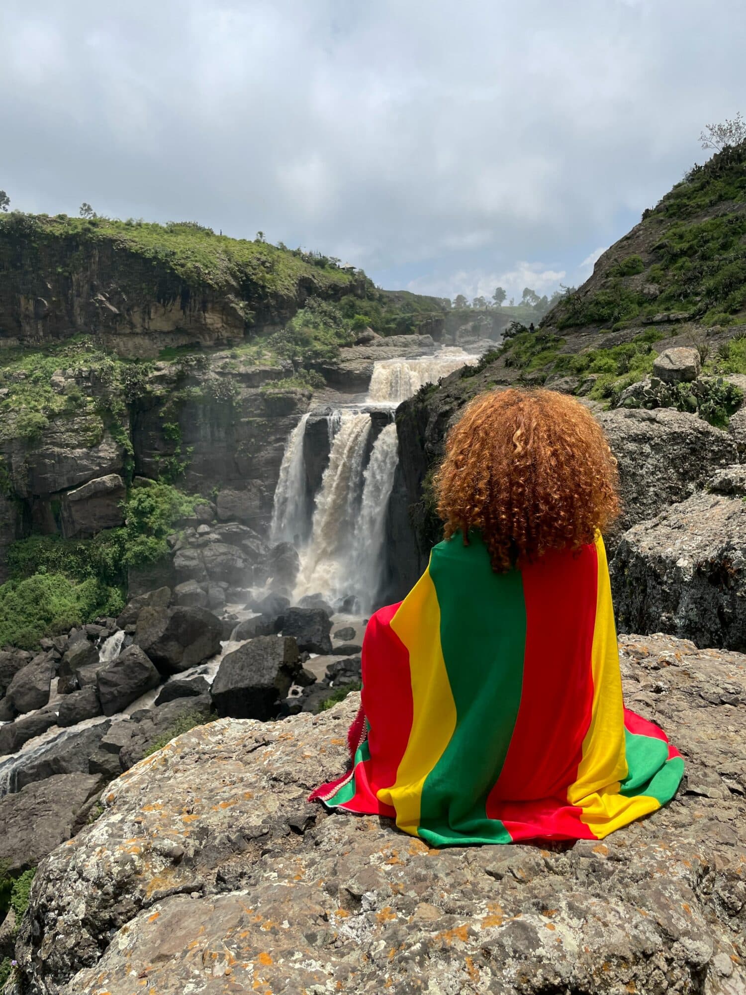 uma mulher de costas embrulhada em uma bandeira com as cores da Etiópia, sentada em frente a uma cachoeira em um ambiente bem rochoso, mas com vegetação, abaixo há o Mosteiro de Debre Libanos, para ilustrar o post de chip celular Etiópia