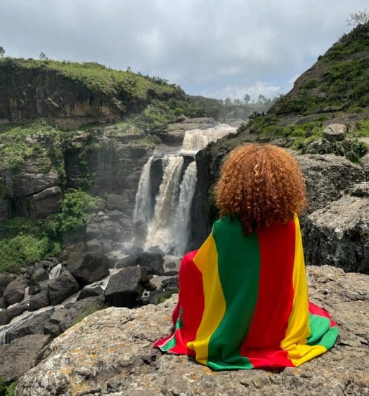 uma mulher de costas embrulhada em uma bandeira com as cores da Etiópia, sentada em frente a uma cachoeira em um ambiente bem rochoso, mas com vegetação, abaixo há o Mosteiro de Debre Libanos, para ilustrar o post de chip celular Etiópia