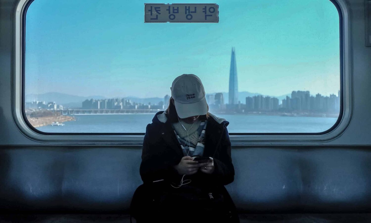 mulher de boné mexendo no celular dentro de um trem no bairro Gangnam-gu, em Seul, com um grande vidro mostrando a cidade moderna ao fundo, com um prédio bem alto em destaque, para ilustrar o post de chip celular Coréia do Sul