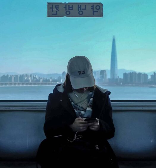 mulher de boné mexendo no celular dentro de um trem no bairro Gangnam-gu, em Seul, com um grande vidro mostrando a cidade moderna ao fundo, com um prédio bem alto em destaque, para ilustrar o post de chip celular Coréia do Sul