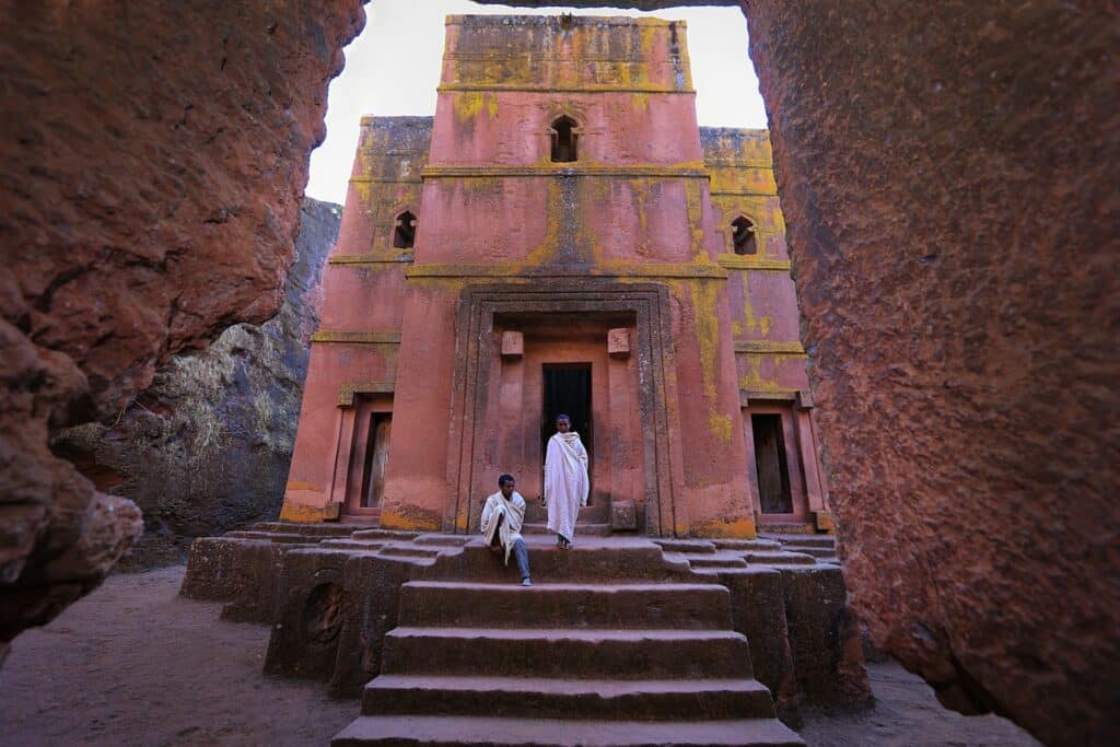 duas pessoas com roupas brancas típicas do país, acima de uma escadaria e na frente de uma igreja esculpida em pedra com torres retas e janelas pequenas, para ilustrar o post de chip celular Etiópia