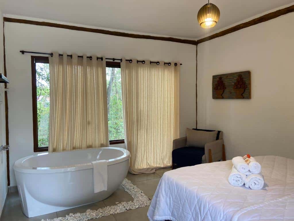 Quarto da A Mi Manera Bistro, com uma cama de casal com toalhas enroladas em cima, uma poltrona cinza e uma banheira de hidro com janela de vidro grande ao lado com algumas cortinas e vista para a natureza verde localizada no lado de fora