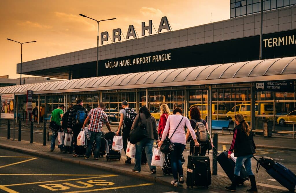 Foto do Aeroporto de Praga, Václav Havel, na República Tchega, pelo lado de fora, com uma fila de pessoas chegando com suas malas e sacolas