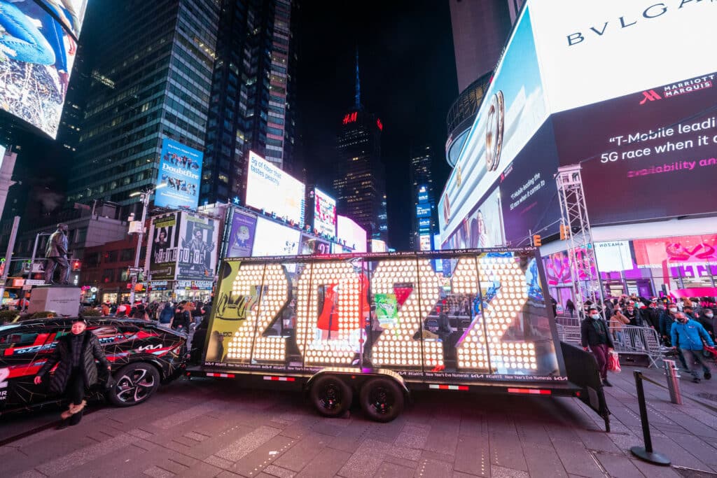 Carro com letreiro iluminado escrito "2022", em região da Times Square, em Nova York, para o ano novo na cidade, com outdoors ao fundo e pessoas andando