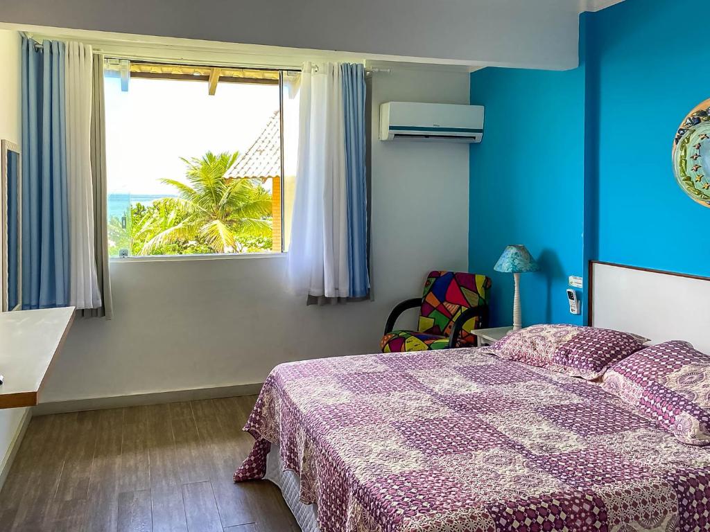 Apartamento deluxe do O Costão do Sol, de 60 m², sendo na imagem uma cama de casal, uma poltrona ao lado com desenhos coloridos e uma mesinha com abajur azul em cima. No lado esquerdo há uma janela com vista das folhas de uma Palmeira e o mar no fundo