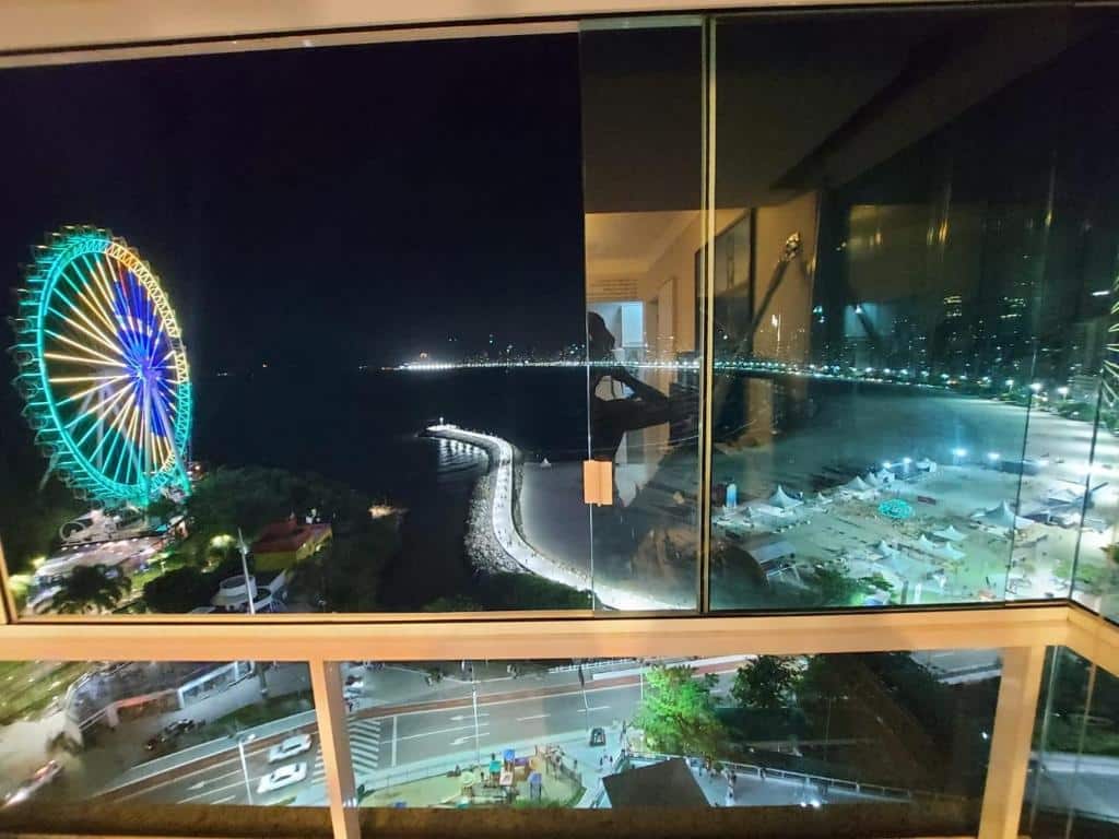 Janela de vidro na sacada do Apto top, a melhor vista de Balneário Camboriú!, com vista da praia em uma noite iluminada pelas luzes da cidade e uma roda gigante iluminada com o desenho da bandeira do Brasil