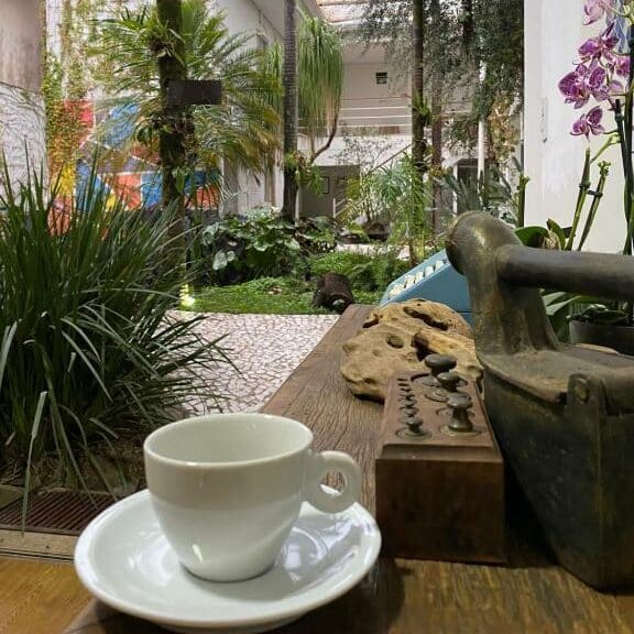 Vista da área de descanso do Hotel e Pousada Oca com jardim e mesa com uma xícara de café branca. Representa hotéis em Holambra.
