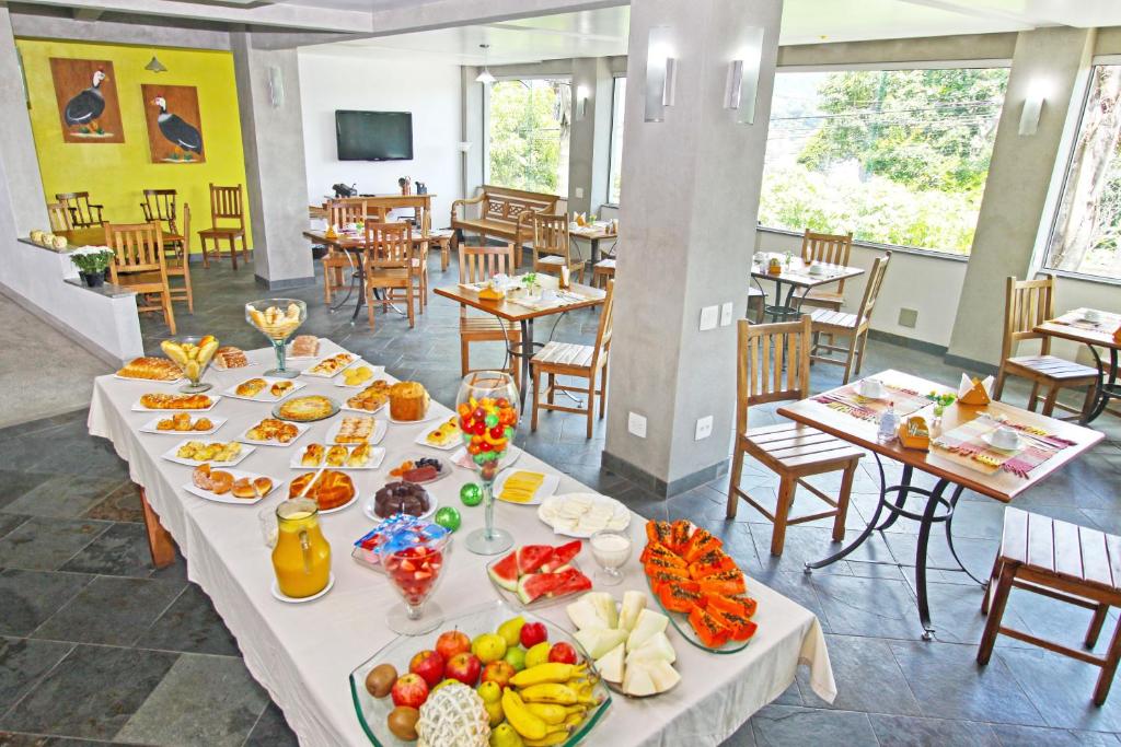 Café da manhã da hospedagem Aventureiro pousada em Teresópolis com frutas, pães, e suco em uma mesa grande e outras mesas menores em volta.