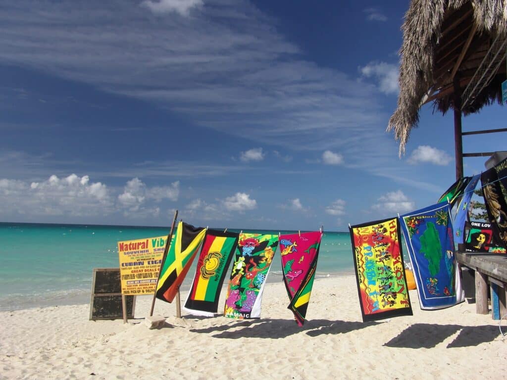 Toalhas coloridas e estampadas penduradas em um varal numa praia para ilustrar o post sobre chip de celular para a Jamaica. A areia é branca, o mar tem água cristalina e o céu é azul com poucas nuvens. Atrás do varal há uma placa amarela com escritos em inglês. - Foto: via Pxhere