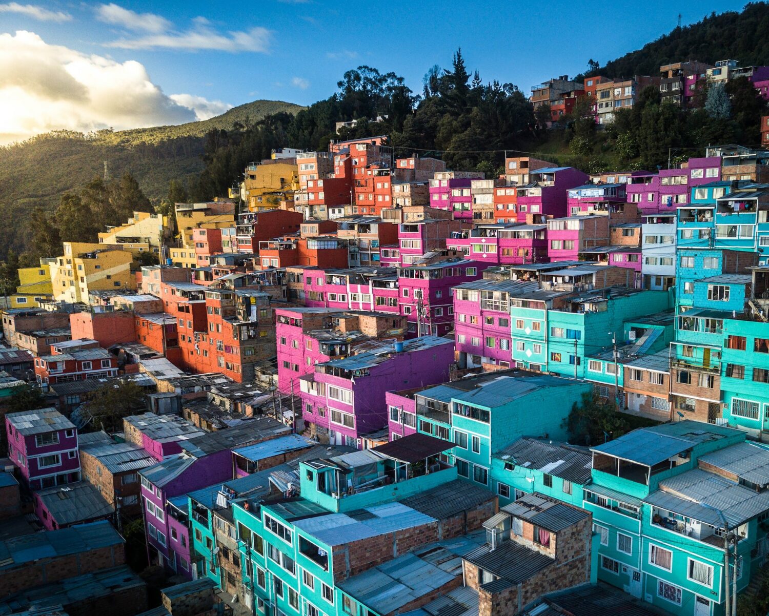 Diversas casas das cores azul, rosa, laranja e amarelo ao longo de uma colina. Do lado esquerdo podemos ver algumas nuvens acima de vegetação, e o céu é azul. - Foto: Enrique Hoyos via Pexels