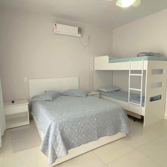 Suíte da Bouganville Guest House, de 20 m², com uma cama de casal, uma beliche e uma mesinha branca ao lado de cada cama. As paredes e o piso são brancos e há um ar-condicionado na parede