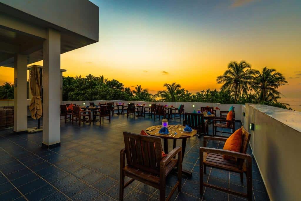 Área externa di Boutique Beach All Inclusive Diving Hotel, em Maldivas, com mesas e cadeiras espalhadas, com árovres em volta e o céu mostrando um pôr do sol laranja
