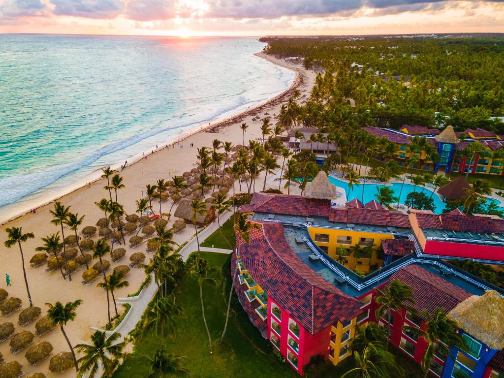 Vista do Caribe Deluxe Princess, localizado à beira-mar, com prédios coloridos nas cores amarelo, pink e azul, piscinas, bastante natureza em volta e espreguiçadeiras espalhas na areia da praia, que tem águas azul claro. O céu ponta um fim de tarde, com um raio de sol mais alaranjado
