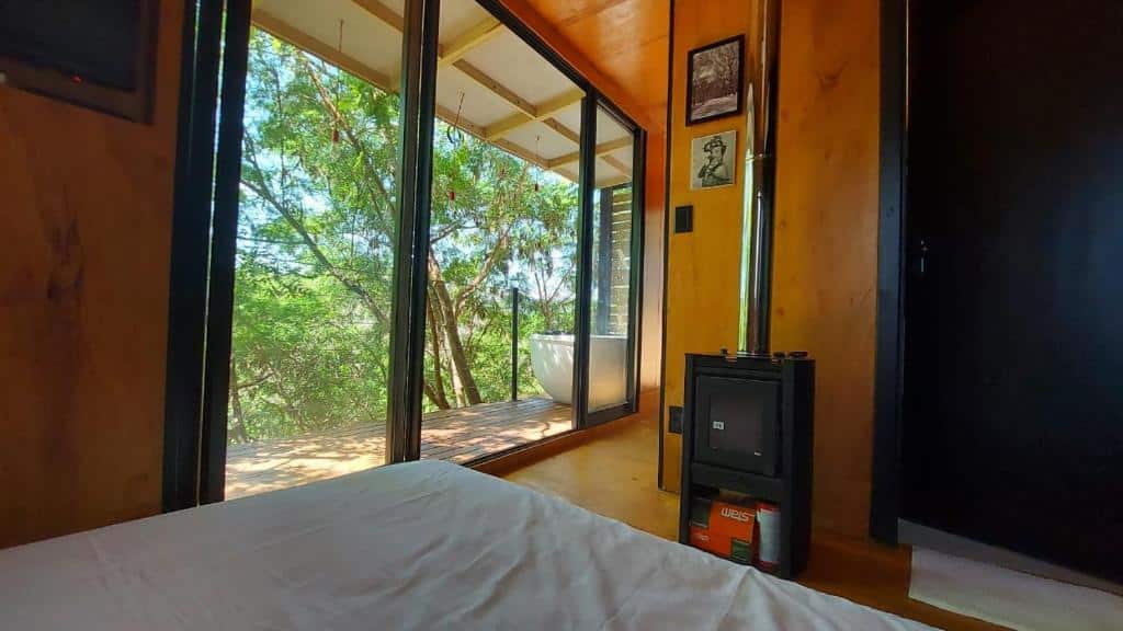 Interior da Casa da Árvore espaço Vila da Serra, com uma cama de casal, lareira e porta grande de vidro dando acesso à uma hidro na varanda com vista para a natureza