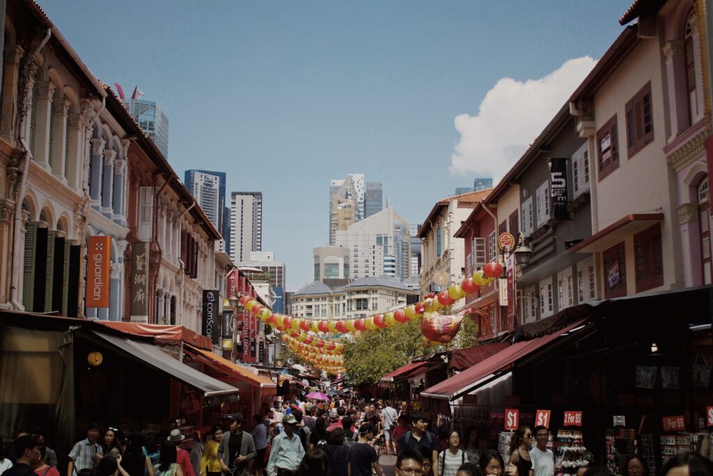 Multidão de pessoas passando por uma rua do bairro Chinatown, uma das recomendações de onde ficar em Singapura. Ao fundo é possível ver alguns prédios com um céu azul acima. - Foto: K8 via Unsplash